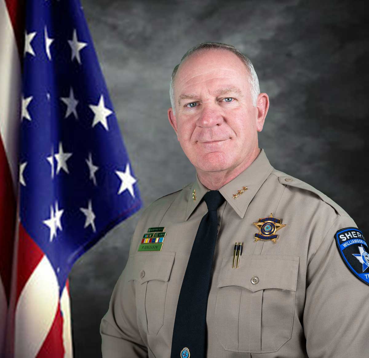 Chief Deputy Patrick Erickson standing next to an American Flag on a textured grey background